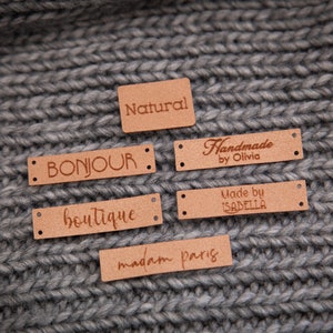 May include: Six brown leather labels with different text on each. The labels are on a gray knitted fabric background. The text on the labels reads: "Natural", "Bonjour", "Boutique", "Handmade by Olivia", "Made by Isabella", and "madam paris".