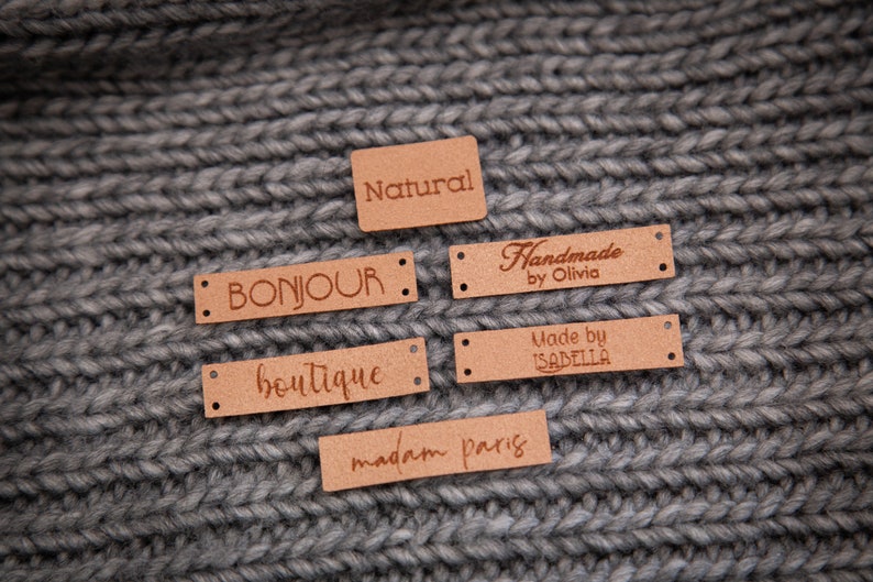 May include: Six brown leather labels with different text on each. The labels are on a gray knitted fabric background. The text on the labels reads: "Natural", "Bonjour", "Boutique", "Handmade by Olivia", "Made by Isabella", and "madam paris".