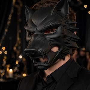 Pode incluir: Uma máscara de lobo preta fosca com detalhes de pelo esculpidos, orelhas pontiagudas e uma boca aberta com dentes visíveis. A máscara cobre a parte superior do rosto, revelando os olhos, e é usada com uma camisa preta de colarinho.