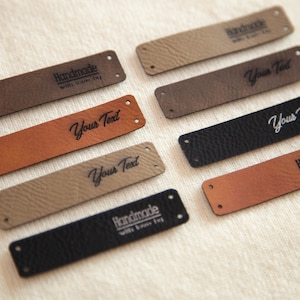 May include: An assortment of rectangular leather tags in shades of brown and black. Some tags are inscribed with "Handmade with love by", others with "Your Text". Each tag is pre-drilled with two holes for attachment.