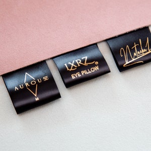 Può includere: Tre etichette in raso nero con lettere dorate. La prima etichetta riporta "AUROUS" con un design a triangolo. La seconda etichetta riporta "IXRZ EYE PILLOW". La terza etichetta riporta "Natasha".