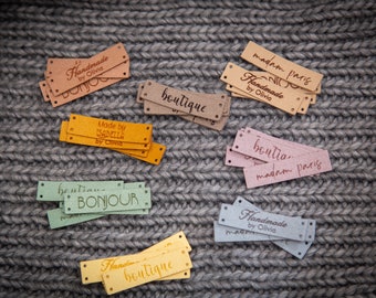 Puede incluir: Una colección de etiquetas rectangulares de piel sintética para ropa en varios colores, incluyendo bronceado, amarillo, verde claro, rosa y gris. Cada etiqueta tiene agujeros preperforados en los extremos y texto personalizado como Handmade by Olivia.
