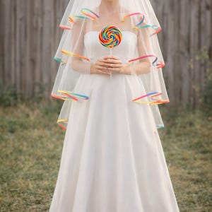 Op de afbeelding: Een bruid in een witte strapless trouwjurk en sluier, met een grote, kleurrijke lolly in haar handen. De sluier is afgezet met regenboogkleurige linten. Ze draagt gele hakken.