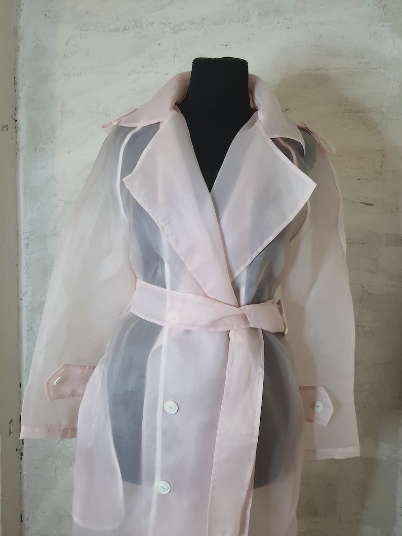 May include: A sheer pink trench coat with a belt and buttons. The coat is made of a lightweight, translucent fabric.