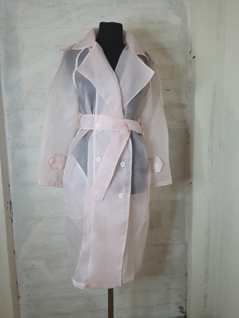 May include: A sheer pink trench coat with a double breasted closure and a belt. The coat is made of a lightweight, translucent fabric.