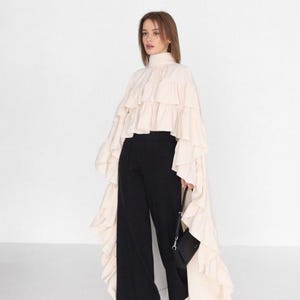 May include: A cream-coloured, high-neck blouse with multiple ruffled layers, paired with black wide-leg trousers. A small black handbag is held in the hand. The outfit is set against a plain white background.