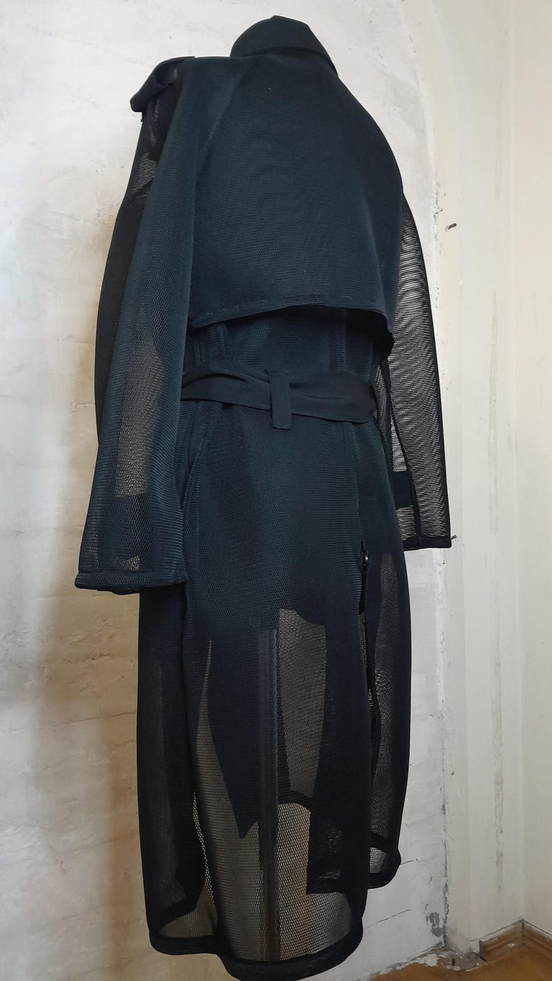 May include: A black mesh trench coat with a belt. The coat is see-through and has a long, flowing silhouette.