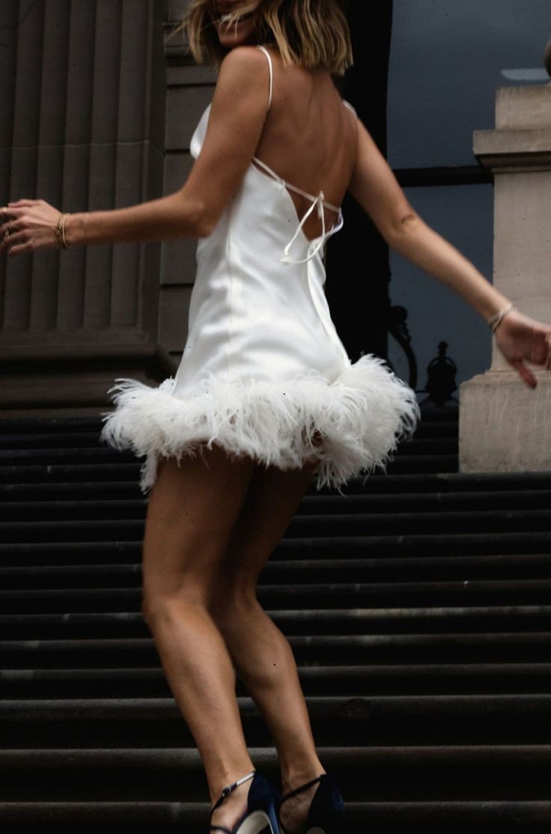May include: A white mini dress with a feather trim. The dress has a cowl neck and a spaghetti strap design. The dress is tied at the back with a bow.