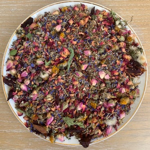 Op de afbeelding: Een witte schaal met een gouden rand gevuld met een kleurrijke mix van gedroogde bloemen en kruiden. De mix bevat rozenblaadjes, lavendel, korenbloemen en andere botanische ingredi&euml;nten. De opstelling is een levendige mix van roze, paars, blauw en groen.
