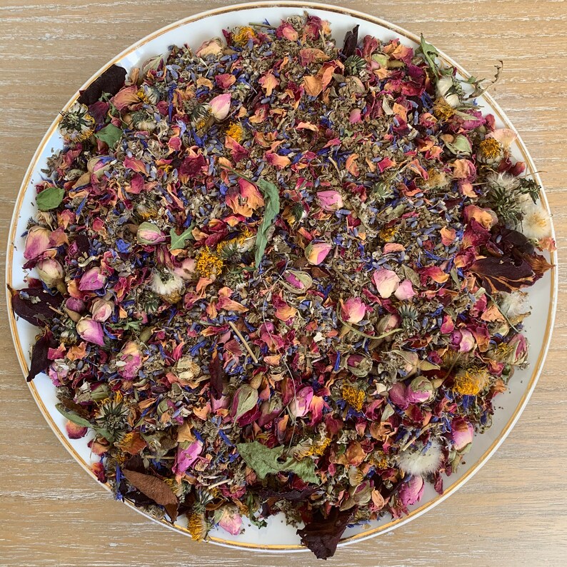Op de afbeelding: Een witte schaal met een gouden rand gevuld met een kleurrijke mix van gedroogde bloemen en kruiden. De mix bevat rozenblaadjes, lavendel, korenbloemen en andere botanische ingredi&euml;nten. De opstelling is een levendige mix van roze, paars, blauw en groen.