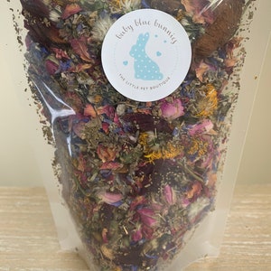 Op de afbeelding: Een doorzichtige plastic zak gevuld met gedroogde bloemen en kruiden. De mix bevat roze rozenblaadjes, blauwe korenbloemen en gele bloemen. Een ronde sticker met een blauw konijn en de tekst "baby blue bunnies".