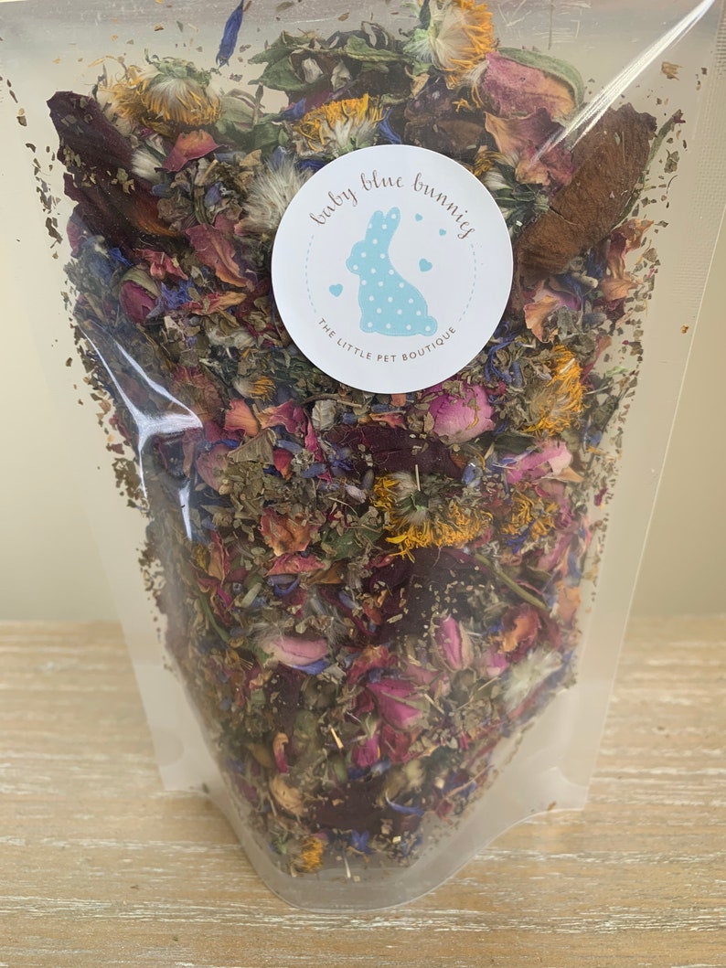 Op de afbeelding: Een doorzichtige plastic zak gevuld met gedroogde bloemen en kruiden. De mix bevat roze rozenblaadjes, blauwe korenbloemen en gele bloemen. Een ronde sticker met een blauw konijn en de tekst "baby blue bunnies".