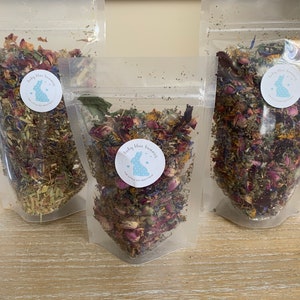Op de afbeelding: Drie transparante zakjes gevuld met gedroogde bloemen en kruiden. Elk zakje heeft een ronde sticker met een blauw konijnenlogo en tekst. De inhoud omvat roze rozenblaadjes, blauwe korenbloemen en groene kruiden.