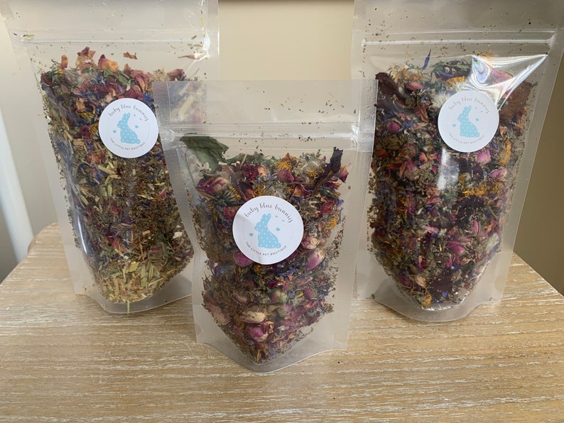 Op de afbeelding: Drie transparante zakjes gevuld met gedroogde bloemen en kruiden. Elk zakje heeft een ronde sticker met een blauw konijnenlogo en tekst. De inhoud omvat roze rozenblaadjes, blauwe korenbloemen en groene kruiden.