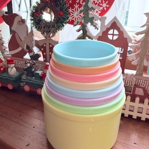May include: A set of pastel-coloured nesting bowls. The bowls are in shades of yellow, green, blue, pink, and orange. The bowls are made of plastic and designed to fit inside each other for storage. The background includes Christmas decorations.