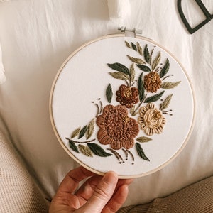 Peut inclure: Un cercle de broderie blanc avec un motif floral composé de fleurs brunes, vertes et jaunes. Les fleurs sont brodées sur un fond de tissu blanc.