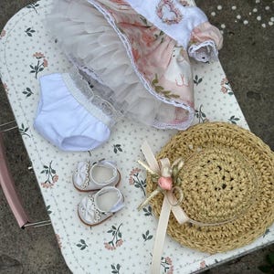 May include: A doll's outfit set, including a floral dress with a tulle skirt, white bloomers, and matching shoes with bows. A straw hat with a ribbon and flower accents completes the ensemble. The items are arranged on a floral patterned surface.