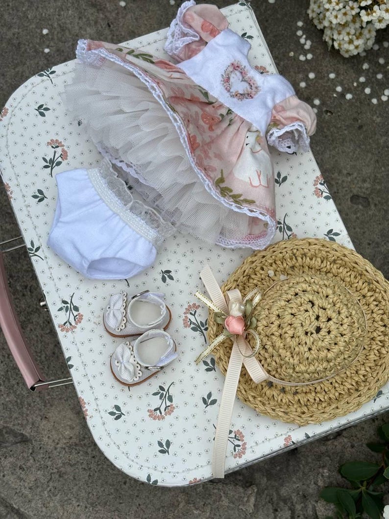 May include: A doll's outfit set, including a floral dress with a tulle skirt, white bloomers, and matching shoes with bows. A straw hat with a ribbon and flower accents completes the ensemble. The items are arranged on a floral patterned surface.