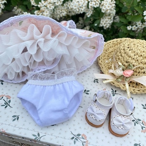 May include: A doll's outfit featuring a pink and white floral dress with a ruffled skirt, white bloomers, a straw hat with a pink ribbon and flower, and white shoes with bows. The items are arranged on a floral patterned surface.
