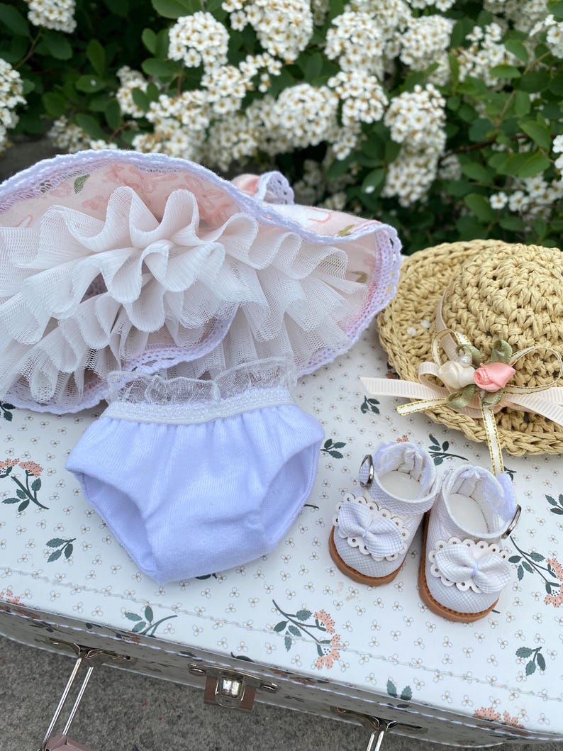 May include: A doll's outfit featuring a pink and white floral dress with a ruffled skirt, white bloomers, a straw hat with a pink ribbon and flower, and white shoes with bows. The items are arranged on a floral patterned surface.
