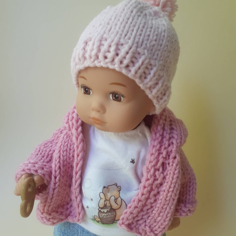May include: A pink knitted hat with a pom pom on top, worn by a doll with a pink knitted cardigan and a white t-shirt with a Winnie the Pooh illustration. The doll is holding a small wooden toy.