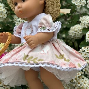 May include: A doll wearing a straw hat, a white top with a floral heart design, and a pink dress with a swan pattern. The dress has white lace trim. The doll is wearing white shoes with a buckle.