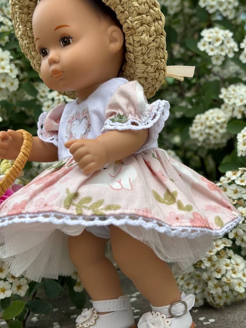 May include: A doll wearing a straw hat, a white top with a floral heart design, and a pink dress with a swan pattern. The dress has white lace trim. The doll is wearing white shoes with a buckle.