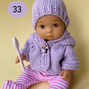 May include: A purple knitted doll outfit with a matching hat and a white striped pair of pants. The doll is holding a white spoon.