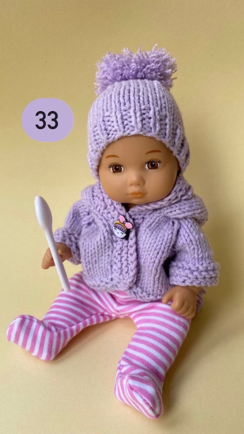 May include: A purple knitted doll outfit with a matching hat and a white striped pair of pants. The doll is holding a white spoon.