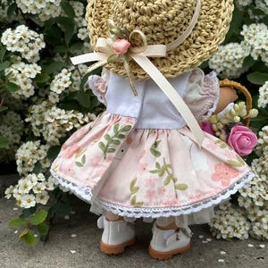 May include: A doll wearing a straw hat with a pink ribbon and a floral dress. The dress is light pink with a white lace trim. The doll is also wearing white shoes with brown soles and carrying a basket of flowers.