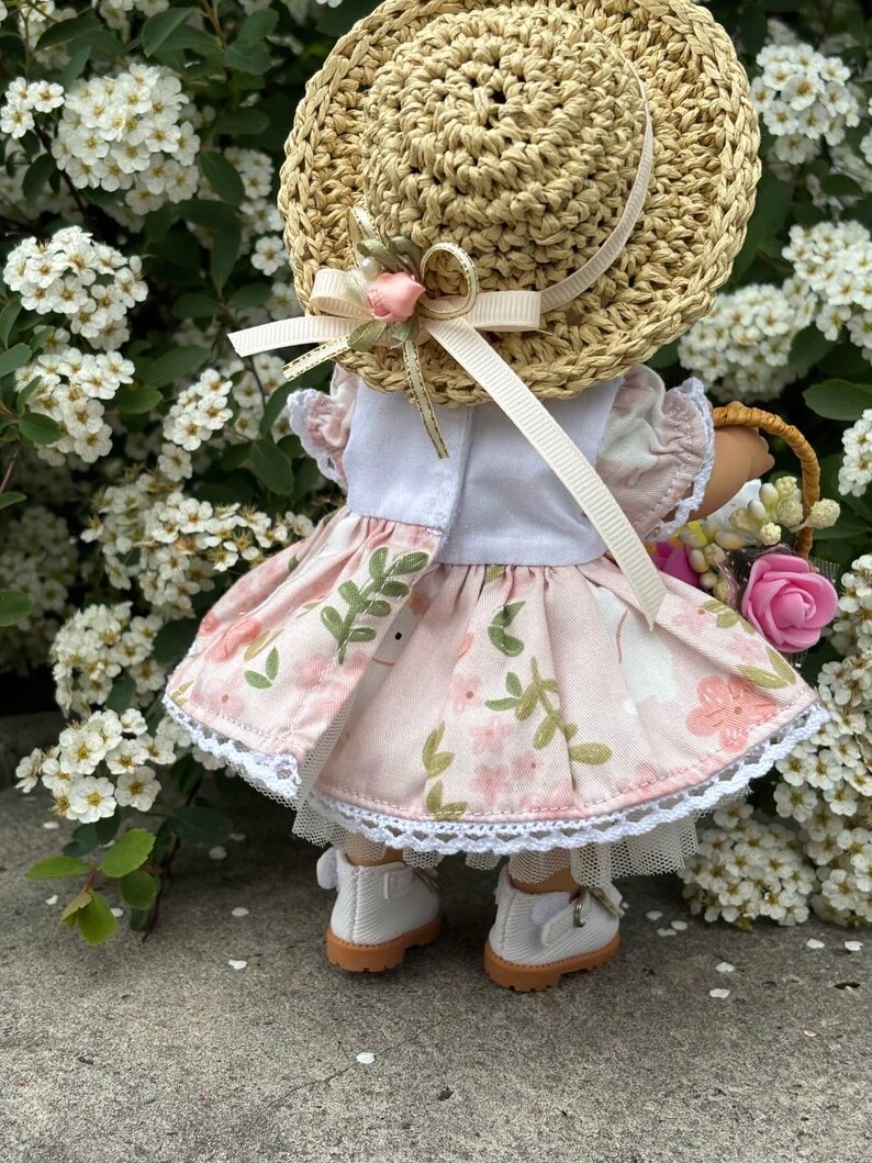 May include: A doll wearing a straw hat with a pink ribbon and a floral dress. The dress is light pink with a white lace trim. The doll is also wearing white shoes with brown soles and carrying a basket of flowers.