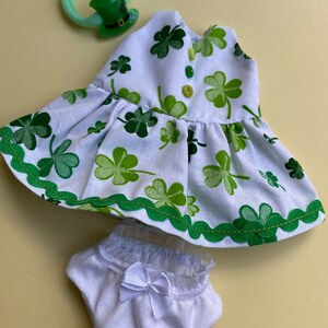 May include: A white dress with a green shamrock print, a white diaper with a white bow, and a green shamrock hair clip.