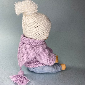 May include: A knitted doll wearing a white hat with a pom-pom, a lavender knitted hooded sweater with a tassel, and blue jeans.