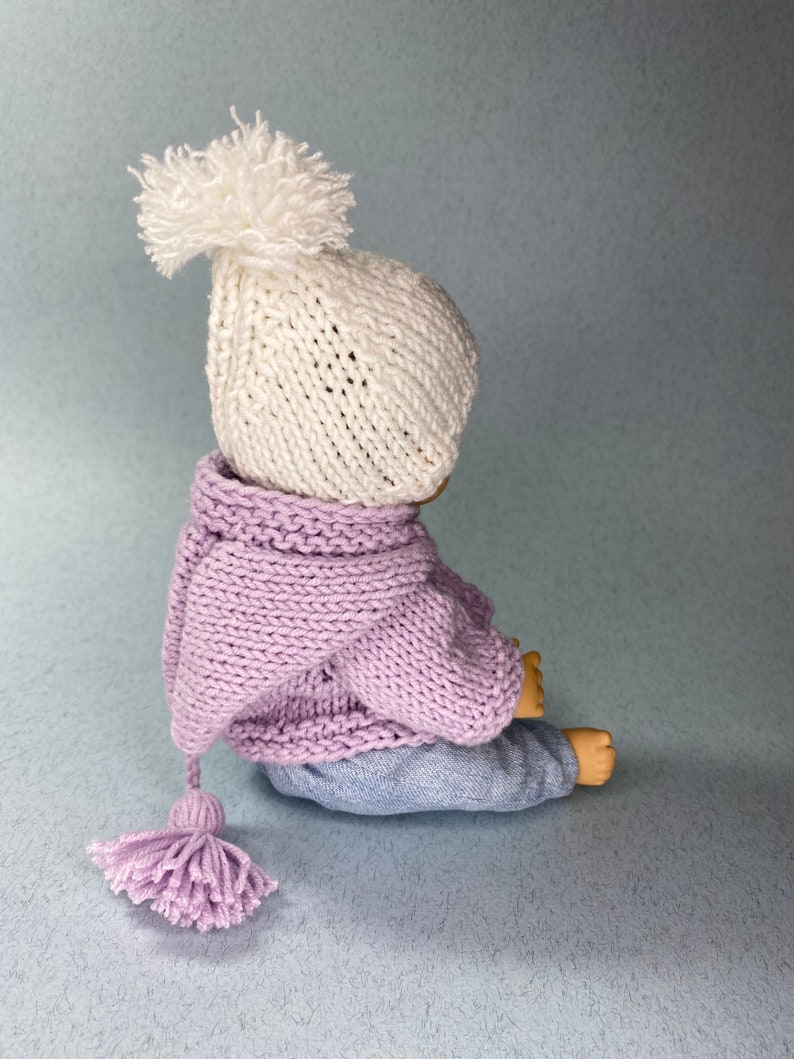 May include: A knitted doll wearing a white hat with a pom-pom, a lavender knitted hooded sweater with a tassel, and blue jeans.
