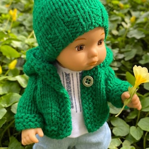 May include: A green knitted hooded jacket with a matching green pom-pom hat. The jacket has a button closure and the doll is holding a yellow flower.