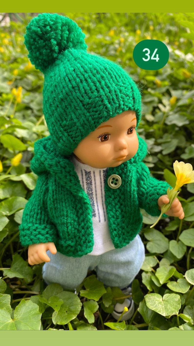 May include: A green knitted hooded jacket with a matching green pom-pom hat. The jacket has a button closure and the doll is holding a yellow flower.