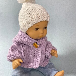 May include: A doll wearing a lavender knitted cardigan with a bee button, white knitted hat with a pom pom, and blue jeans.