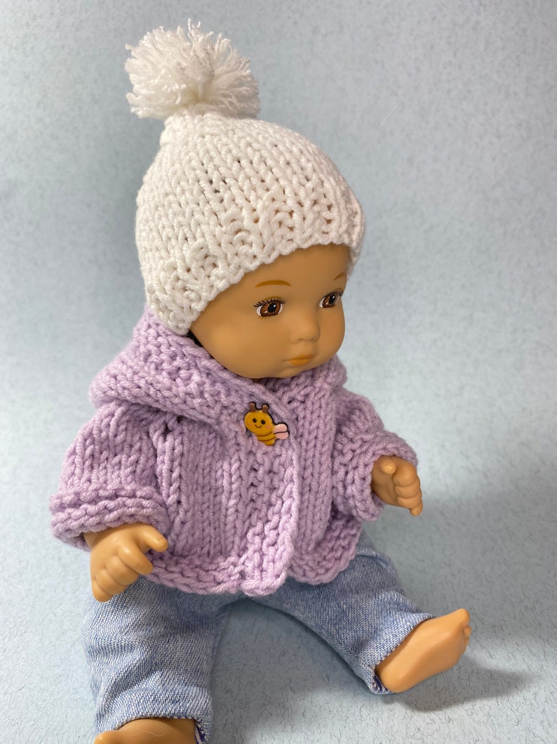 May include: A doll wearing a lavender knitted cardigan with a bee button, white knitted hat with a pom pom, and blue jeans.
