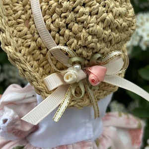 May include: A straw bonnet with a decorative bow featuring a pearl, pink rose, and white ribbon. The bonnet has a light pink ribbon and is part of a floral dress. The bonnet is made of woven straw.