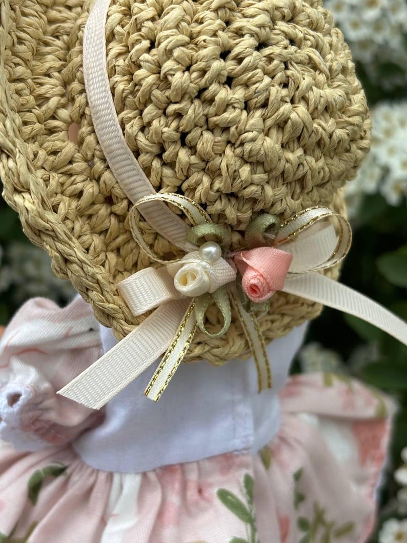 May include: A straw bonnet with a decorative bow featuring a pearl, pink rose, and white ribbon. The bonnet has a light pink ribbon and is part of a floral dress. The bonnet is made of woven straw.