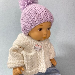 May include: A doll wearing a lavender knitted hat with a pom-pom and a white knitted cardigan with a cat button. The doll is wearing blue jeans and has brown eyes.