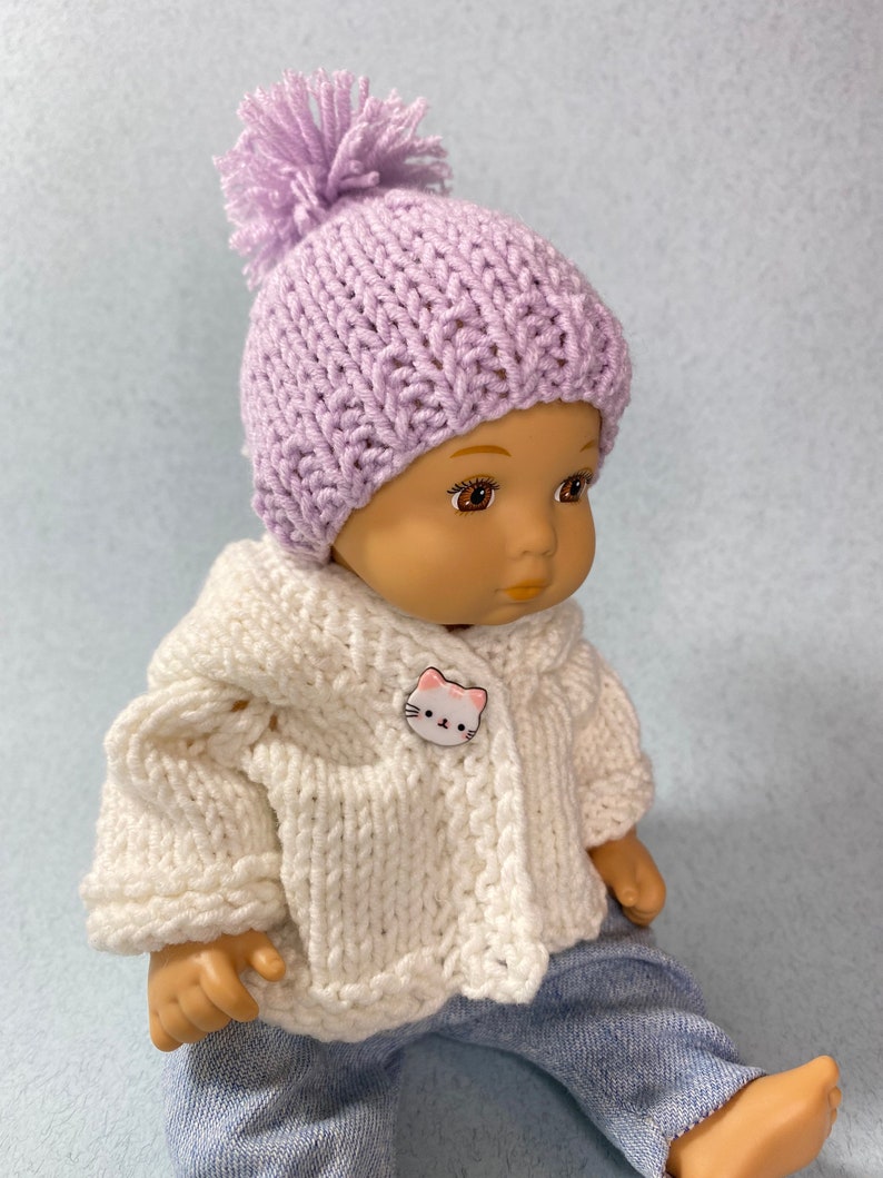 May include: A doll wearing a lavender knitted hat with a pom-pom and a white knitted cardigan with a cat button. The doll is wearing blue jeans and has brown eyes.
