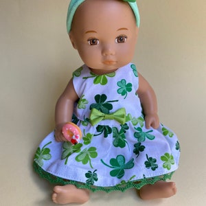 May include: A doll wearing a white dress with green shamrock print, a green headband with a green leprechaun hat, and holding a pink and white donut.