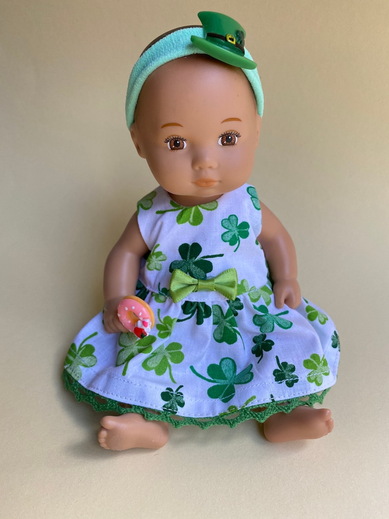 May include: A doll wearing a white dress with green shamrock print, a green headband with a green leprechaun hat, and holding a pink and white donut.