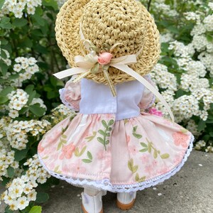 May include: A doll wearing a straw hat with a pink floral dress. The hat has a ribbon and a small pink rose. The dress has a white bodice and a skirt with a floral pattern and white lace trim. The doll is wearing white shoes.