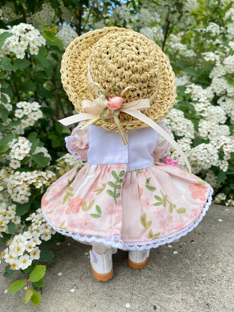 May include: A doll wearing a straw hat with a pink floral dress. The hat has a ribbon and a small pink rose. The dress has a white bodice and a skirt with a floral pattern and white lace trim. The doll is wearing white shoes.