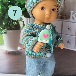 Puede incluir: Una mu&ntilde;eca con un gorro de punto azul y verde, un c&aacute;rdigan de punto azul y verde, pantalones vaqueros azules y zapatillas verdes. La mu&ntilde;eca sostiene una paleta de hielo verde con un coraz&oacute;n rosa.