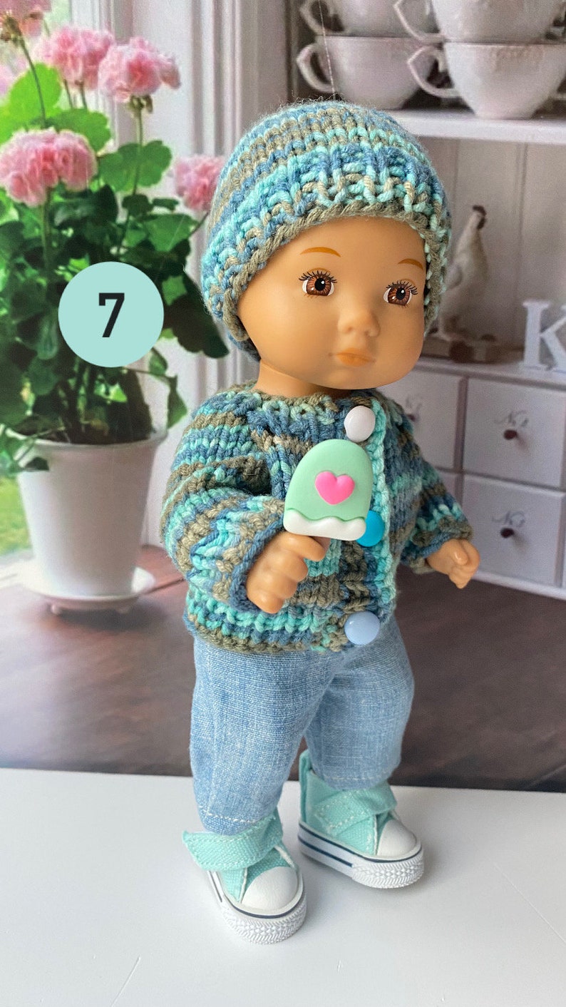 Puede incluir: Una mu&ntilde;eca con un gorro de punto azul y verde, un c&aacute;rdigan de punto azul y verde, pantalones vaqueros azules y zapatillas verdes. La mu&ntilde;eca sostiene una paleta de hielo verde con un coraz&oacute;n rosa.