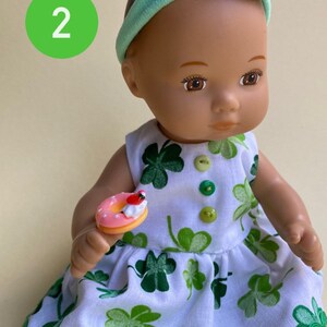 May include: A doll wearing a white dress with green shamrock print, a green headband with a green hat, and holding a pink and white donut. The doll has brown eyes and brown hair.