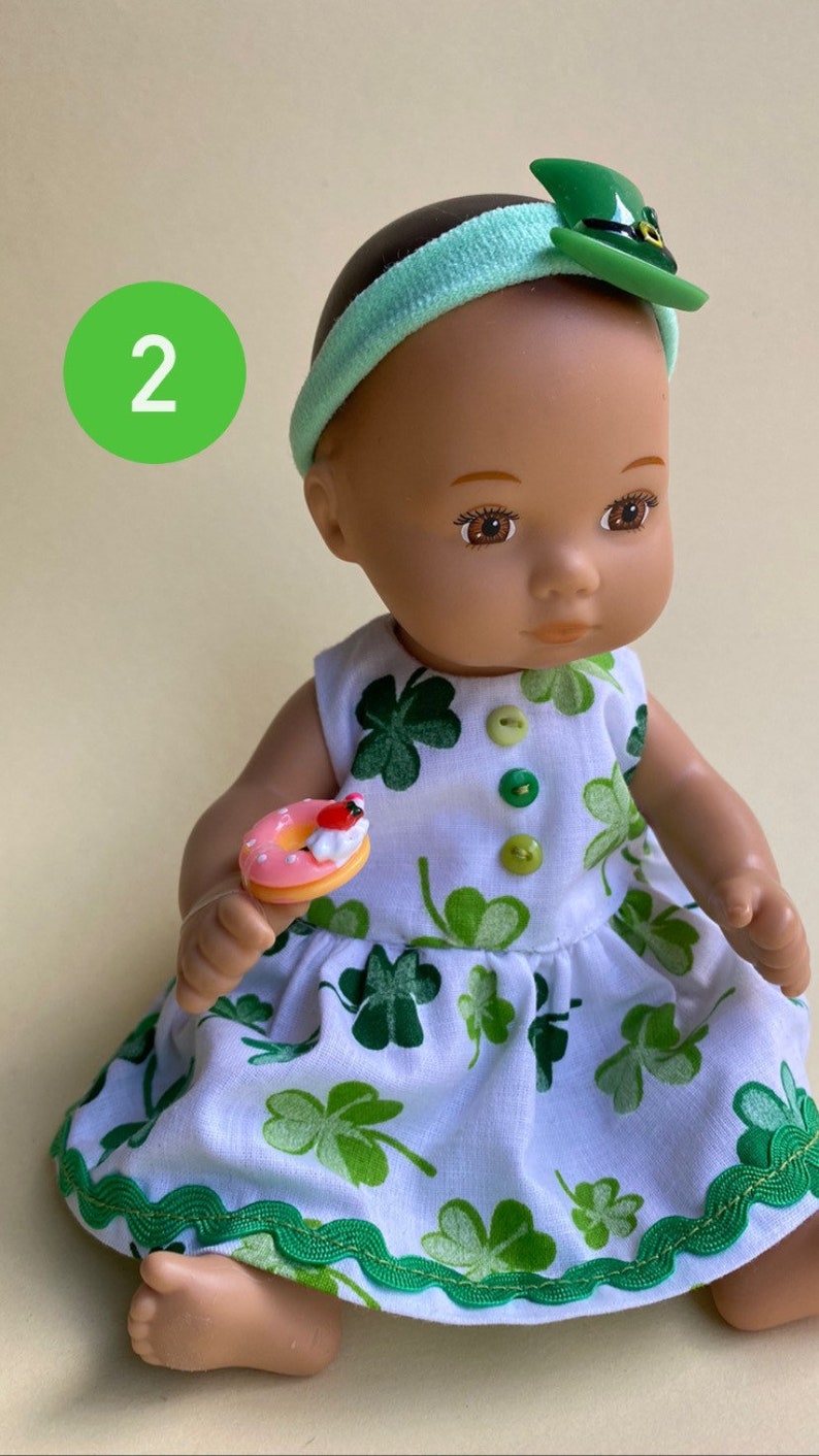 May include: A doll wearing a white dress with green shamrock print, a green headband with a green hat, and holding a pink and white donut. The doll has brown eyes and brown hair.